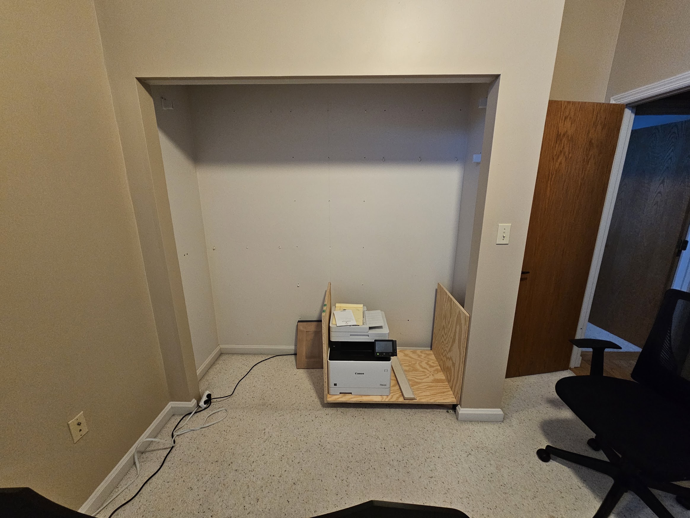 Gutted closet opening with a large printer on the floor beside a rough plywood mock-up
