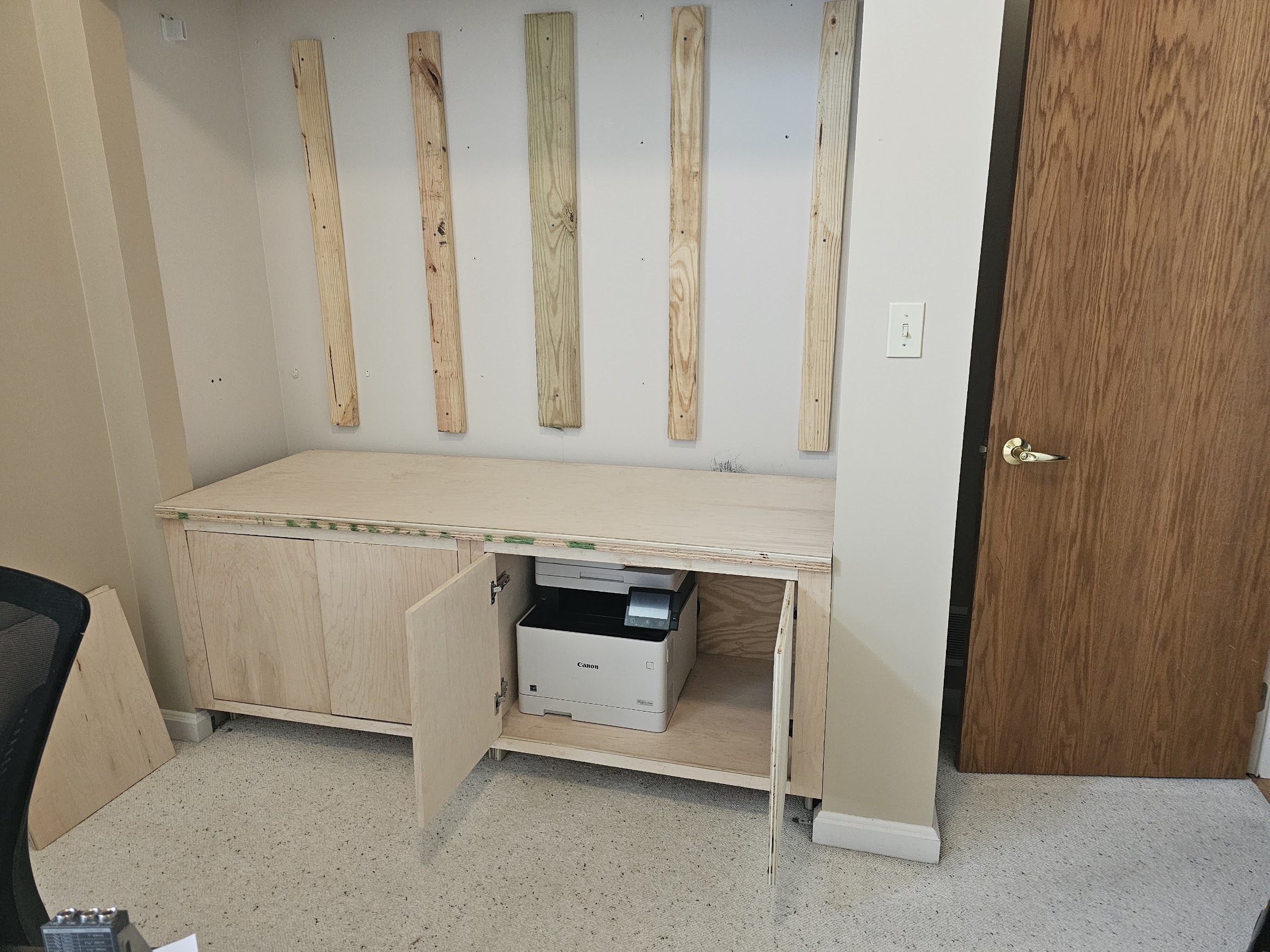 Printer sitting inside the base cabinet with doors open, vertical furring strips mounted on the wall above